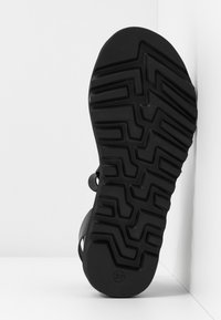 Sandale noire avec une semelle en caoutchouc texturée et un motif en zigzag, bout arrondi et sangles réglables sur les côtés. Taille 37 visible.