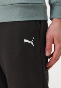 Spodnie dresowe Puma w ciemnym brązowym kolorze z bawełny, z białym logo na udzie. Miękka tkanina o luźnym kroju, z ściągaczem w pasie i u dołu nogawek.