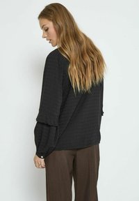 Sort mønstret bluse med flæser på ærmerne, designet med løs pasform. Sammen med brune bukser, som fremhæver en tekstureret stofdetalje.