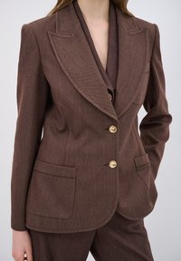 Blazer marrone su misura con rever a sottosquadro, bottoni dorati e tasche laterali. Tessuto testurizzato con un motivo sottile, design aderente.