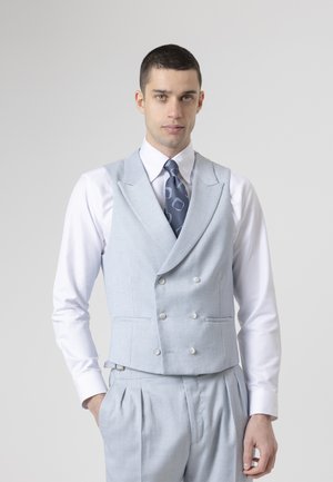 Hombre con chaleco cruzado azul claro, pantalones plisados a juego, camisa blanca y corbata azul con estampado, de pie con una mano en el bolsillo.