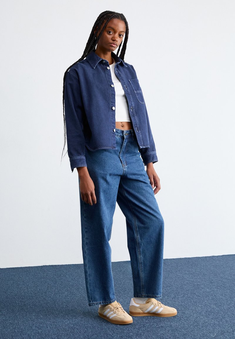 Chaqueta de mezclilla navy, top blanco corto, jeans de tiro alto azul claro y zapatillas beige con franjas blancas. Cabello trenzado y fondo liso.
