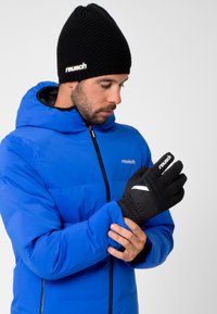 Gants noirs avec une finition texturée et des accents blancs, portés avec une veste rembourrée bleue et un bonnet en tricot noir. Présente un ajustement serré au niveau du poignet.