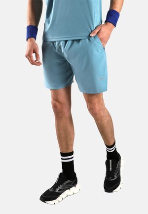 Man draagt lichtblauwe sportshorts, bijpassend shirt, blauwe polsbanden, zwart-wit gestreepte sokken en zwarte hardloopschoenen.