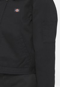 Veste noire à fermeture éclair avec poche avant et petite poche à patch sur la manche, présentant un logo Dickies rectangulaire coloré sur la poitrine.