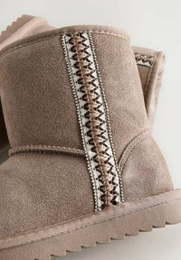 Botines de ante marrón difuso que presentan costuras decorativas en beige y marrón a lo largo del costado, con una suela de goma texturizada.
