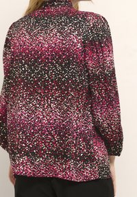 Svart blus med ett gradient prickmönster i rosa, vitt och lila nyanser. Har hög halsringning och puffärmar.