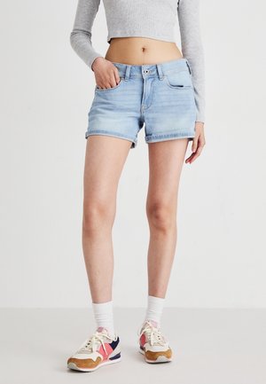 Kvinde iført lyseblå denimshorts, grå langærmet cropped top, hvide sokker og flerfarvede løbesko, står med den ene hånd i lommen.