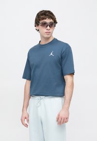 Κοντομάνικο μπλε σκούρο μπλουζάκι με λογότυπο Jordan σε λευκό, συνδυασμένο με ανοιχτό γκρι φόρμα και σκουρόχρωμα γυαλιά ηλίου με γωνιώδη σκελετό.