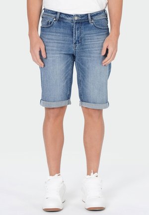 Mann trägt hochgekrempelte blaue Denim-Shorts, weiße Sneaker und ein weißes, in die Hose gestecktes Hemd, steht vor einem schlichten Hintergrund.