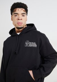 Hoodie noir avec une poche kangourou à l'avant, un capuchon avec cordon de serrage et un logo brodé blanc portant les mots "WORK IN PROGRESS STARGAZER INSTITUTE."