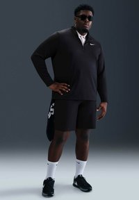 Svart langermet genser med glidelås, kombinert med svarte shorts. Tilbehøret inkluderer hvite sokker og svarte joggesko.