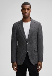 Graue Tweed-Blazer mit subtiler Textur, mit Revers, einreihiger Knopfschluss und seitlichen Taschen. Getragen über einem weißen Hemd.