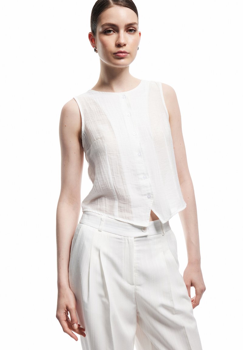 Femme à la peau claire et aux cheveux foncés attachés porte un haut blanc sans manches boutonné et un pantalon blanc plissé taille haute sur fond blanc.