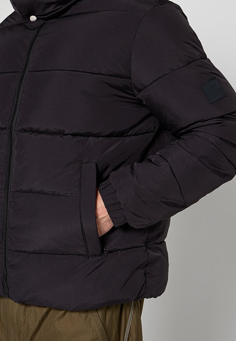 Giacca a vento nera con zip frontale e patch con logo sulla manica, indossata con una mano in tasca laterale, abbinata a pantaloni verdi oliva.