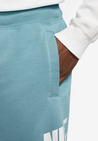 Pantaloni della tuta blu chiaro con texture liscia, dotati di una tasca laterale e dettaglio del logo bianco vicino all'orlo inferiore.