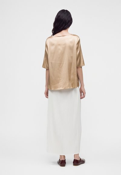 Femme aux longs cheveux foncés portant un haut ample en satin doré, une longue jupe plissée blanche et des chaussures plates marron foncé, debout de dos.