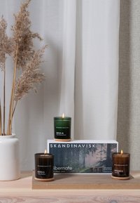 Candele verdi e marroni su un vassoio di legno chiaro, con una scatola bianca etichettata "SKANDENAVISK." Presenta texture morbide e un design minimalista.