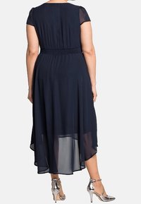 Robe bleu marine à manches courtes, taille smockée, et un voile fluide et transparent. Associée à des chaussures à talons hauts argentées.