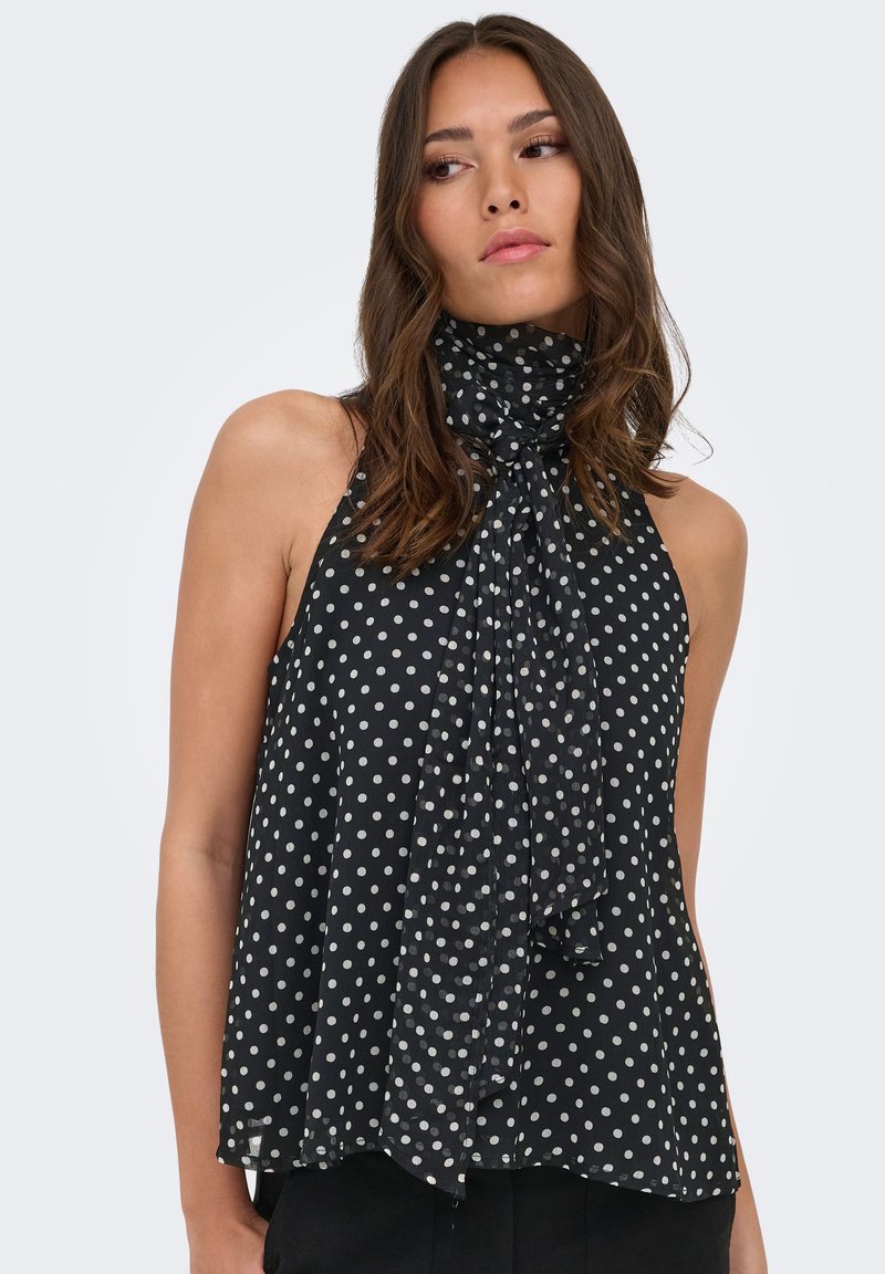 Femme aux longs cheveux bruns portant un haut noir sans manches à pois blancs avec un détail de nœud au col montant, regardant sur le côté.