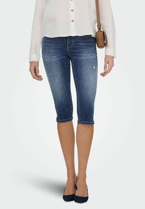 Kvinne iført blå denimshorts til knærne, hvit bluse med lange ermer, svarte hæler og bærer en liten brun skulderveske.