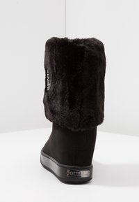 Bota de cuña negra hasta la mitad de la pantorrilla con puño de pelo sintético suave y placa metálica de la marca "GUESS" en el talón, mostrada desde atrás sobre una superficie blanca.