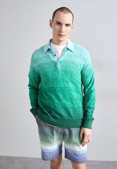 Grüner Farbverlauf-Pullover mit strukturiertem Zickzackmuster, kombiniert mit schwarz-weißen Chevron-Print-Shorts in Blau- und Grüntönen.