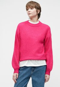 Jauns cilvēks ar īsiem matiem, tērpies spilgti rozā lielā džemperī virs baltas krekla un zila džinsa, pret vienkāršu fona.