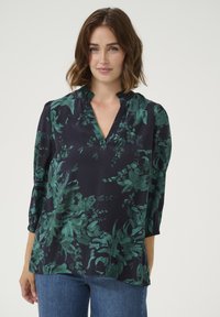 Blouse bleu marine avec un motif floral vert, décolleté en V, détail froncé au niveau du col et manches 3/4. Le tissu semble lisse et léger.