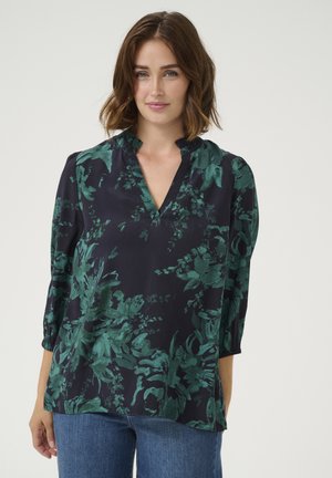 Blouse bleu marine avec un motif floral vert, décolleté en V, détail froncé au niveau du col et manches 3/4. Le tissu semble lisse et léger.