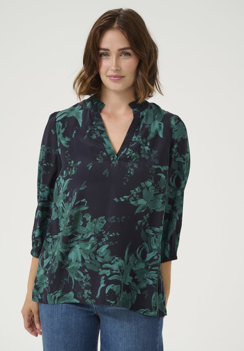 Blouse bleu marine avec un motif floral vert, décolleté en V, détail froncé au niveau du col et manches 3/4. Le tissu semble lisse et léger.