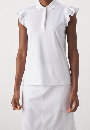 Vrouw die een mouwloze witte polo met gerafelde schouderdetails en een witte rok draagt tegen een effen achtergrond.