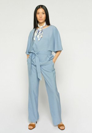 Mujer con mono azul claro con cinturón, pañuelo de seda estampado alrededor del cuello y tacones marrones de punta abierta, de pie con las manos en los bolsillos.