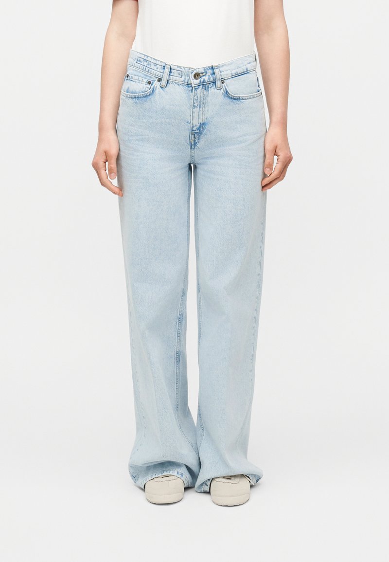 Person som har på seg lyseblå vide jeans og hvite joggesko, stående mot en enkel hvit bakgrunn.