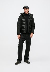 Ung man med svart keps, vadderad svart jacka med huva, svart polotröja, lösa svarta jeans och svarta skor, står framför en vit bakgrund.