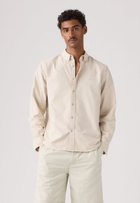 Homme aux cheveux courts et bouclés portant une chemise beige boutonnée et un short de couleur claire, debout les mains dans les poches devant un fond uni.