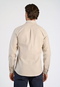Beige_langärmliges Hemd mit klassischem Kragen, Rückenfalten-Detail und geknöpften Manschetten. Der Stoff erscheint glatt mit einer subtilen Textur.