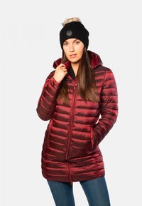 Veste rembourrée rouge avec un design matelassé horizontal, fermeture éclair à l'avant, capuche et poches latérales. Portée avec un bonnet en tricot noir orné d'un pompon en fausse fourrure.
