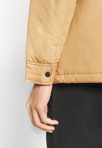 Main en pantalon noir à côté de la manche d'une veste beige avec détail de poignet boutonné, sur un fond clair uni.