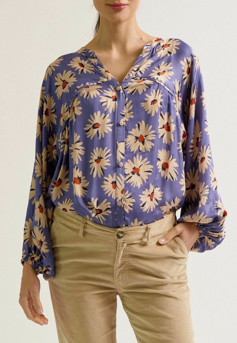 Lila Bluse mit floralen Muster, das cremefarbene Gänseblümchen mit roten Mittelpunkten zeigt. Lange, gepuffte Ärmel und Knopfleiste. Trägt sie mit beigen Hosen.