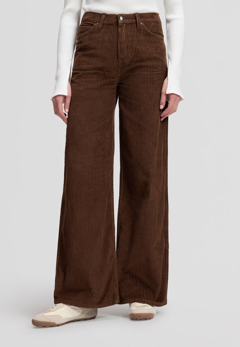 Femme portant un pantalon en velours côtelé marron taille haute, coupe large, associé à des baskets blanches et un haut blanc à manches longues.