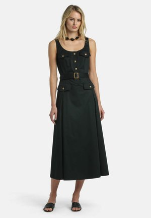 Blonde vrouw die een mouwloze zwarte midi jurk draagt met gouden knopen, riem, voorzakken, zwarte open-toe sandalen en een zwarte ketting.