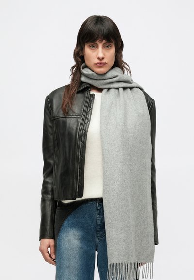 Sciarpa di lana grigia drappeggiata intorno al collo, indossata con una giacca di pelle nera, un maglione color crema e jeans di denim blu, con dettaglio di frange.