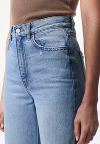 Vaaleansiniset korkeavyötäröiset farkut, jotka on valmistettu denimistä. Ominaisuuksina purjehtivat helmat ja kontrastivärinen ompelu. Napitettu ja vetoketjulausua edessä.