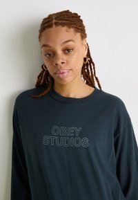 Crna majica dugih rukava s natpisom "OBEY STUDIOS" u bijeloj boji. Ima okrugli izrez i glatku tkaninu. Model ima pletenice.