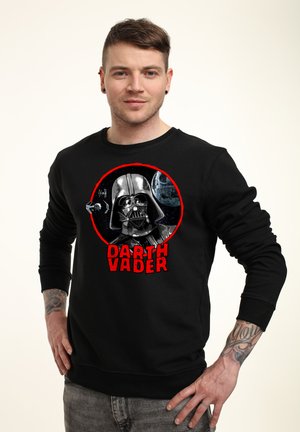 Homme portant un sweat-shirt noir avec casque de Dark Vador, Étoile de la Mort, chasseur TIE et texte rouge « DARTH VADER » sur le devant, debout les mains sur les hanches.