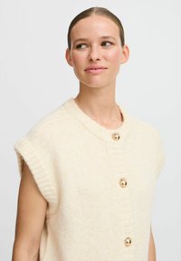 Cremfarbener, kurzärmeliger Strickcardigan mit rundem Ausschnitt, der vorne drei große goldene Knöpfe und eine weiche Textur aufweist.