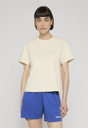 Jeune femme aux cheveux courts et foncés portant un t-shirt beige uni à manches courtes et un short bleu avec un petit logo blanc sur la jambe gauche.