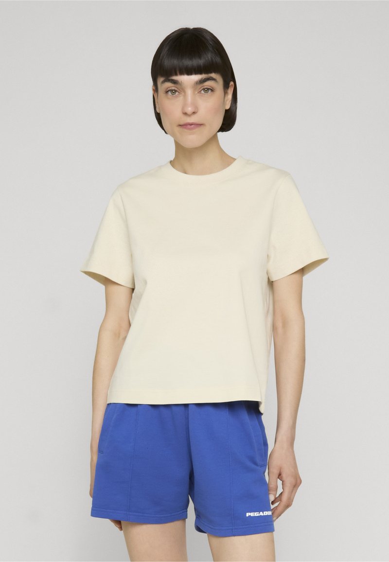 Jeune femme aux cheveux courts et foncés portant un t-shirt beige uni à manches courtes et un short bleu avec un petit logo blanc sur la jambe gauche.