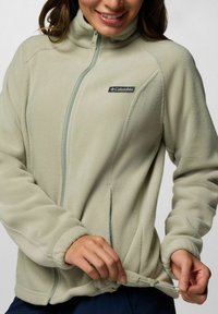 Hellgrüne Fleecejacke mit hohem Kragen, durchgehendem Reißverschluss und verstellbarem Bund. Auf der oberen linken Seite befindet sich das Columbia-Logo. Weicher Stoff.